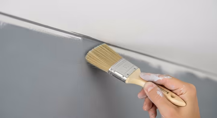 A painter carefully cutting in paint with an angled brush along the top edge of a wall where it meets the ceiling.