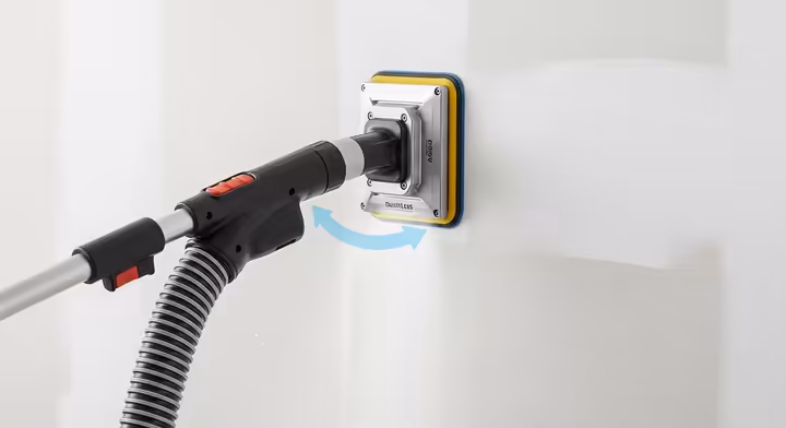 A close-up of a dustless sanding head attached to a HEPA vacuum, being used on a wall to capture dust at the source.
