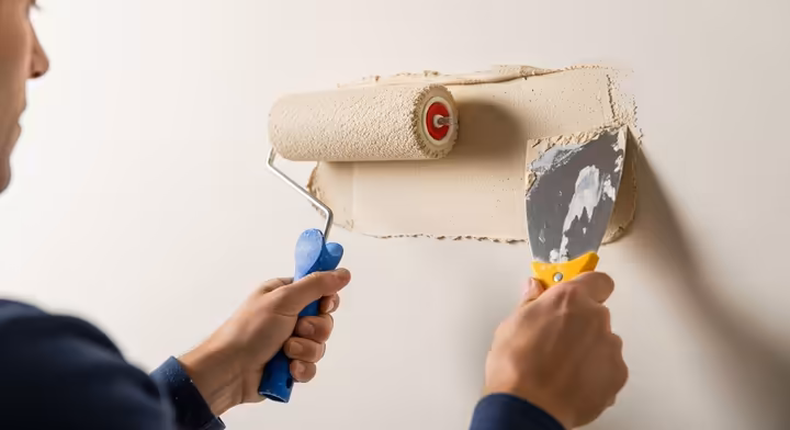 A person applying joint compound to a wall with a paint roller in one hand, while holding a wide taping knife in the other, ready to smooth.